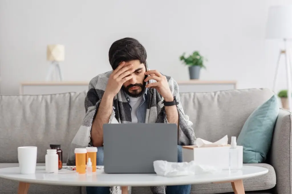 Sick millennial arab man covered in blanket sitting on couch, having conversation on phone while