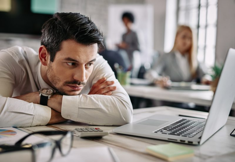 Young businessman leaning on his desk and feeling bored while surfing the net on a computer at work. There are people in the background.